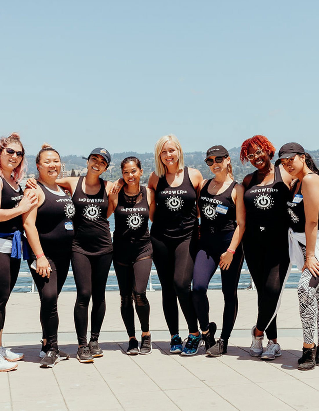 Group of women in black athletic gear standing outdoors on a waterfront promenade, smiling with arms around each other, under a clear blue sky.