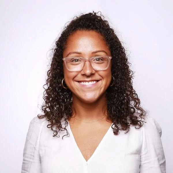 A woman with curly hair wearing glasses and a white top smiling at the camera against a plain background.