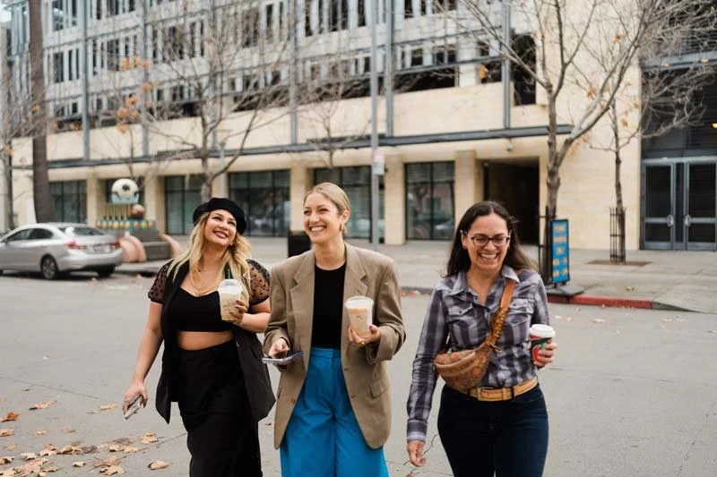 Three young women walking on a city street, smiling and holding coffee cups, with a modern building and parked cars in the background.