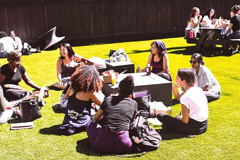 Group of young women sitting on grass in a park, enjoying a sunny day, with tables of food and drinks, and others socializing in the background.