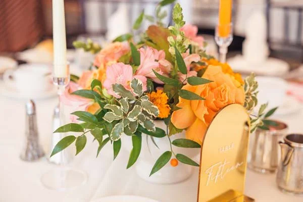 A floral centerpiece with pink, orange, and yellow flowers, surrounded by green leaves, on a white table with candles and silverware.