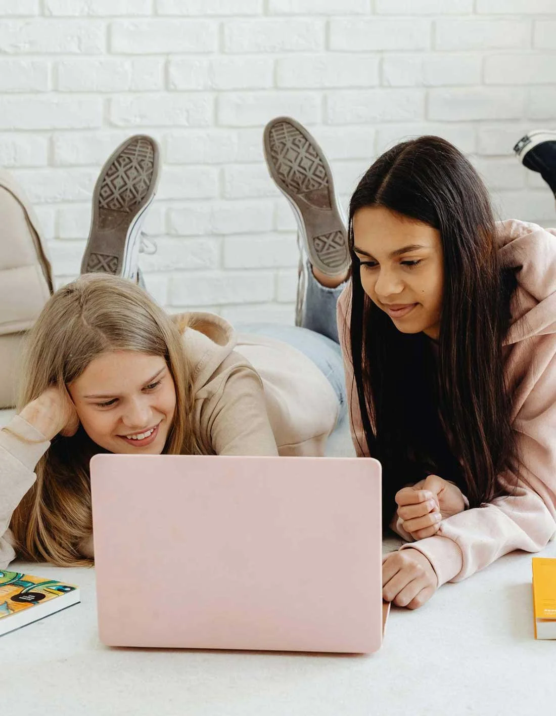 Two young girls watching a laptop screen together, lying on the floor with one girl lying on her stomach and the other sitting beside her.