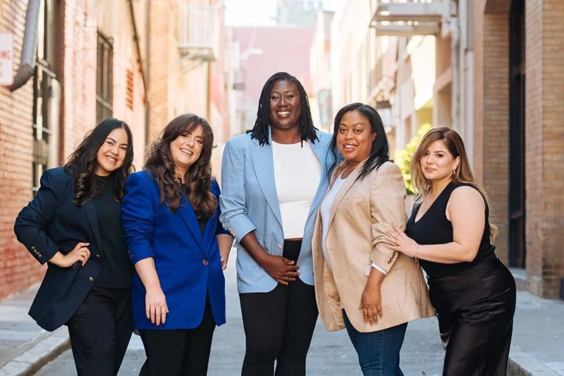 Five women standing together in an urban alleyway, smiling and posing for the photo, dressed in business casual attire.