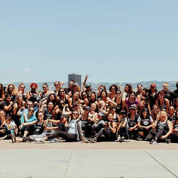 A large group of people posing for a photo outdoors on a sunny day with a cityscape in the background, many wearing black shirts.
