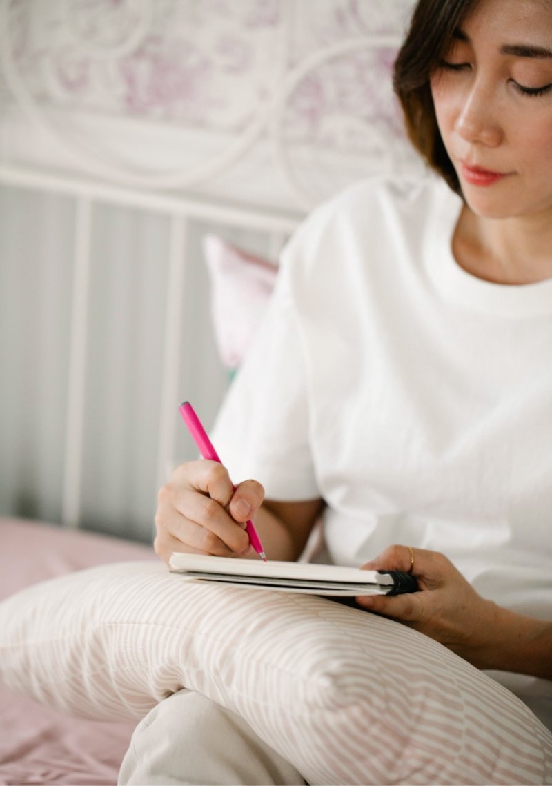Woman sitting on bed, writing in a notebook with a pink pen, in a cozy room with pastel-colored wall and bedding.