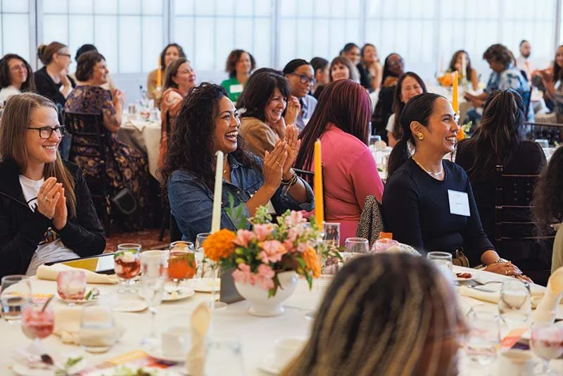 Group of women at an indoor event, some clapping and smiling, seated at round tables decorated with flowers and candles.
