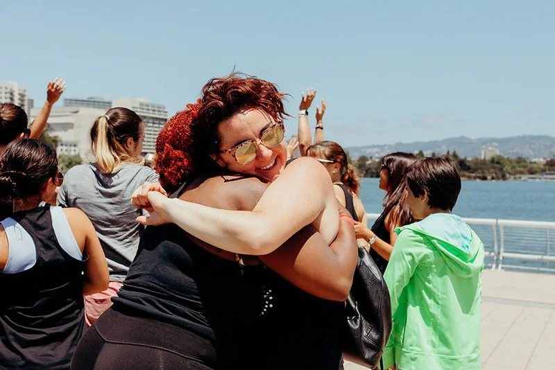 A group of people hugging and smiling outdoors near a body of water on a sunny day.