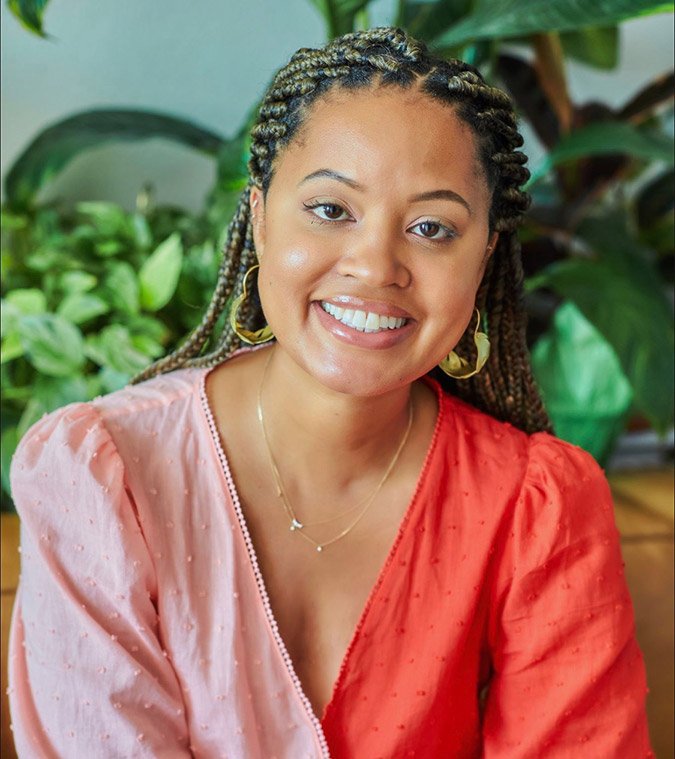 A woman with braided hair, wearing gold hoop earrings and a layered necklace, smiling in front of lush green plants.