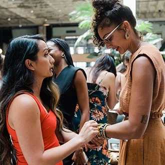 Two women smiling and holding hands in a social gathering at an indoor venue with other people in the background.