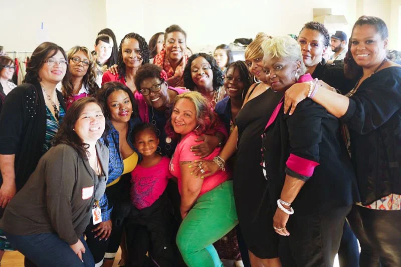 Group of diverse women and girls smiling and gathering together indoors, posing for a photo.