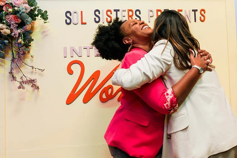 Two women hugging and smiling at an event with a sign in the background that reads "SOL SISTERS PRESENTS INTERNATIONAL WOMEN" and floral decorations.