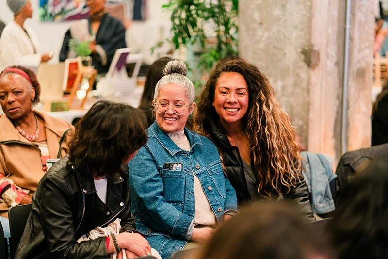 Three women sitting and smiling at an event, with people and plants in the background.