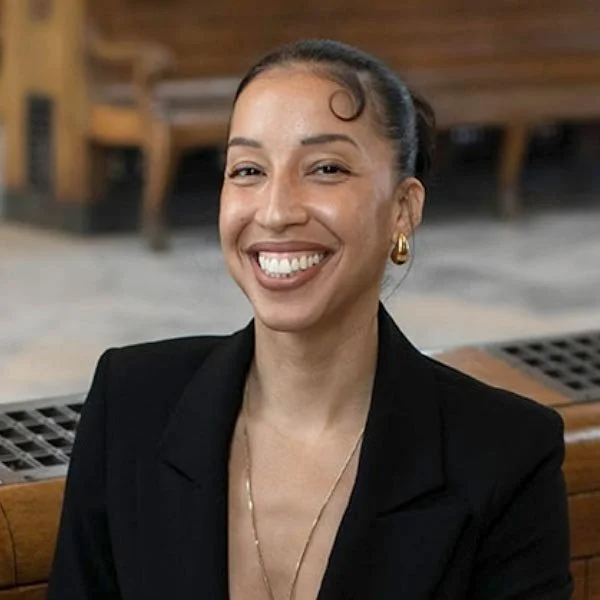 A woman with dark hair styled in a bun, wearing a black blazer and gold jewelry, smiling at the camera in an indoor setting with wooden furniture in the background.