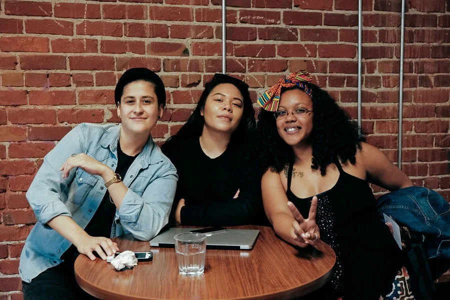 Three women sitting at a wooden table against a brick wall, smiling and posing for the photo.