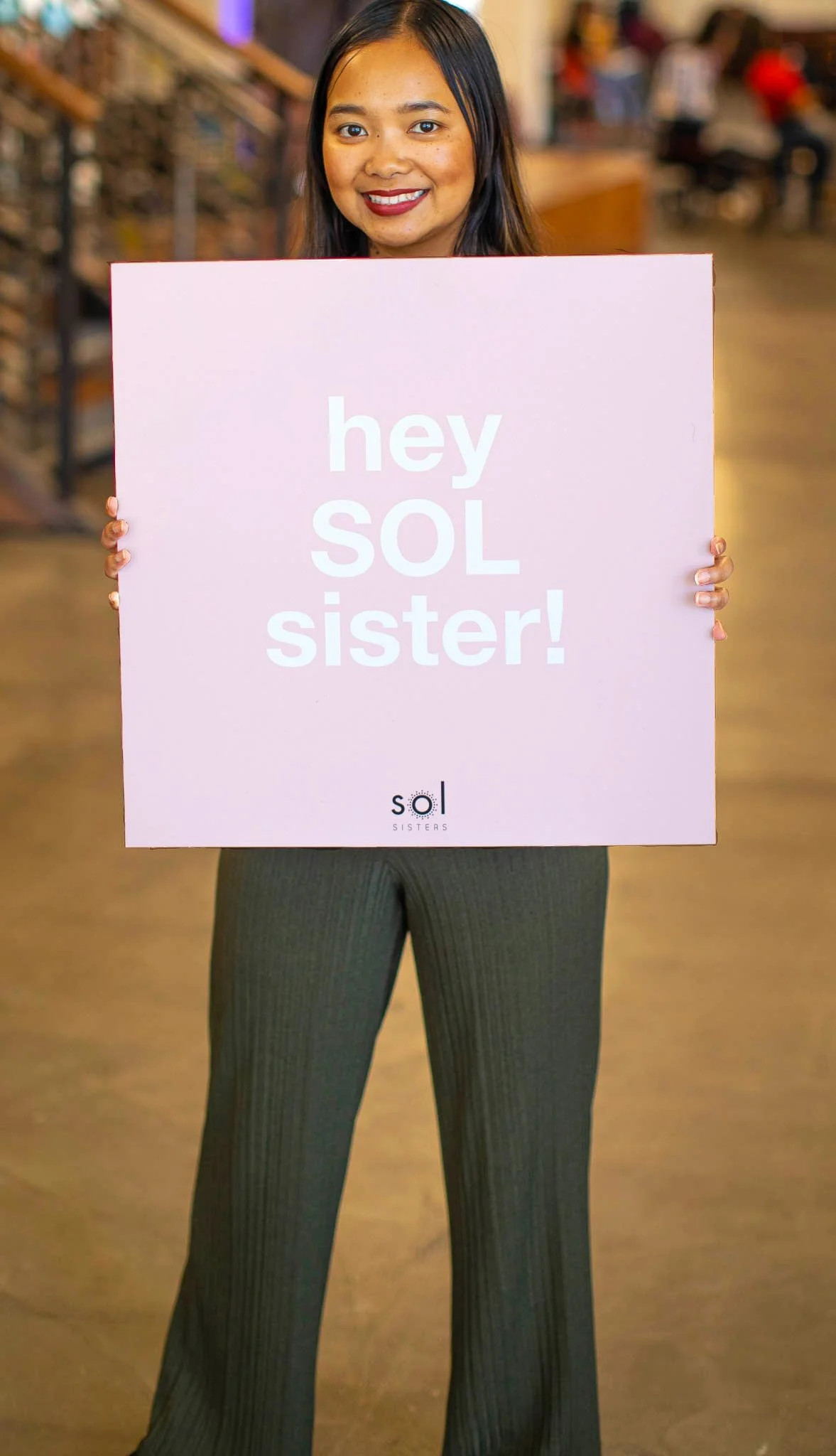 A woman with long dark hair and a warm smile holding a pink sign that says 'hey SOL sister!' with a blurred background of people and indoor setting.