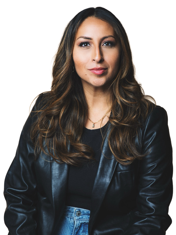 A woman with long wavy brown hair, wearing a black leather jacket over a black shirt, and blue jeans, standing against a neutral background.