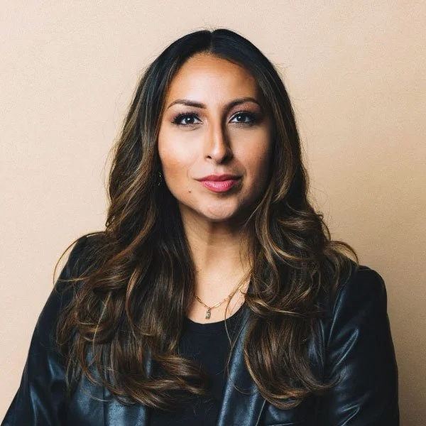 Portrait of a woman with long wavy brown hair, wearing a black leather jacket and a black top, standing against a beige background.