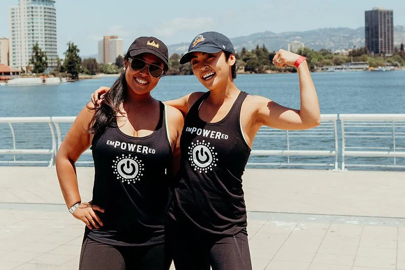 Two women wearing matching black tank tops with white power symbols and the word "EMPOWERED" printed on them, smiling and posing outdoors by a body of water with city buildings in the background.