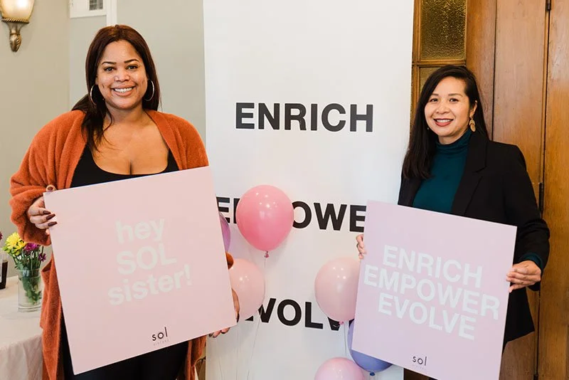 Two women smiling and holding signs that say 'hey SOL sister!' and 'ENRICH EMPOWER EVOLVE' at an event with pink balloons and a white background that says 'ENRICH EMPOWER EVOLVE'.