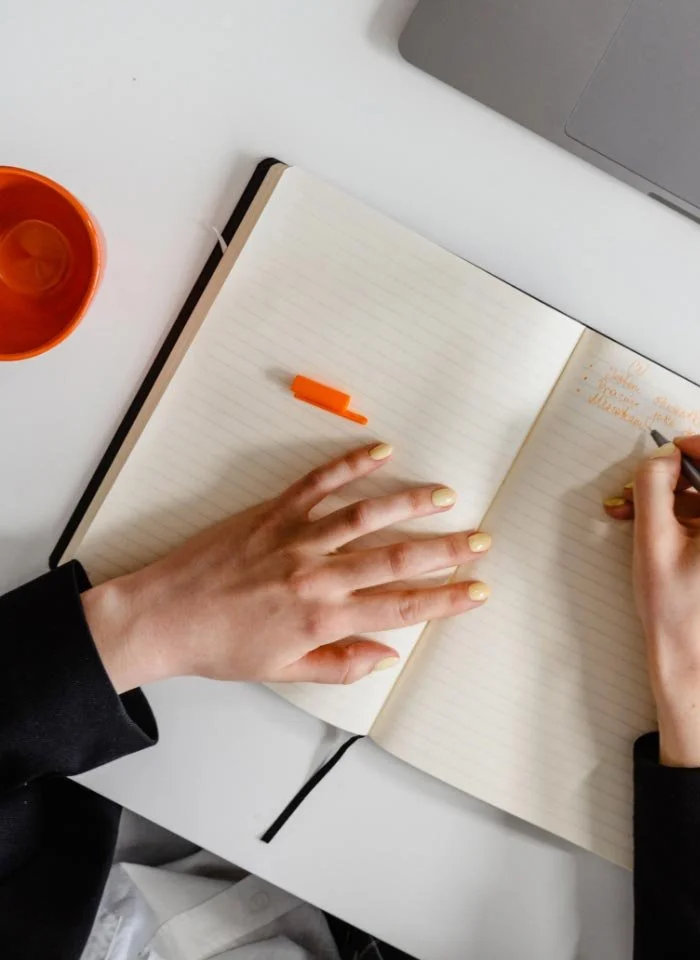 An open notebook with lined pages on a white table, a hand with painted nails is writing in the notebook, with an orange pen cap resting on the notebook. A small orange cup is on the left side and a gray laptop is partially visible at the top.