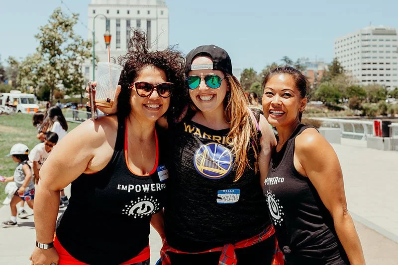 Three women smiling outdoors, wearing black tank tops with the word 'Empowered' on them, at a community event or gathering.