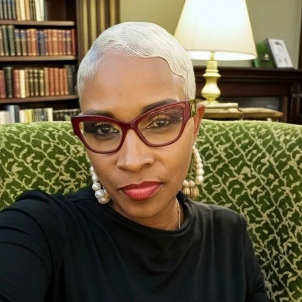 A woman with short, platinum blonde hair and red glasses smiling at the camera. She is wearing pearl earrings and a black top, sitting on a green patterned chair with a bookshelf and a lamp in the background.