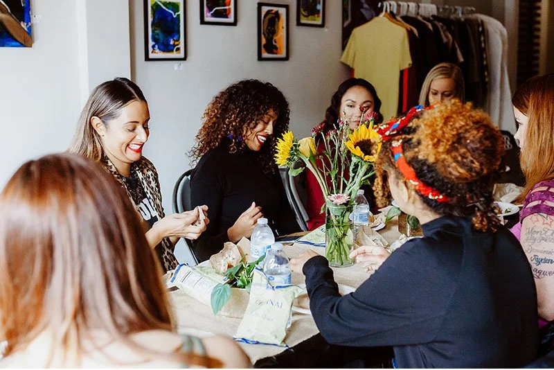 Six women sitting around a table decorated with flowers, water bottles, and paper bags, engaged in conversation and smiling in a cozy indoor setting with artwork on the wall and clothes hanging in the background.