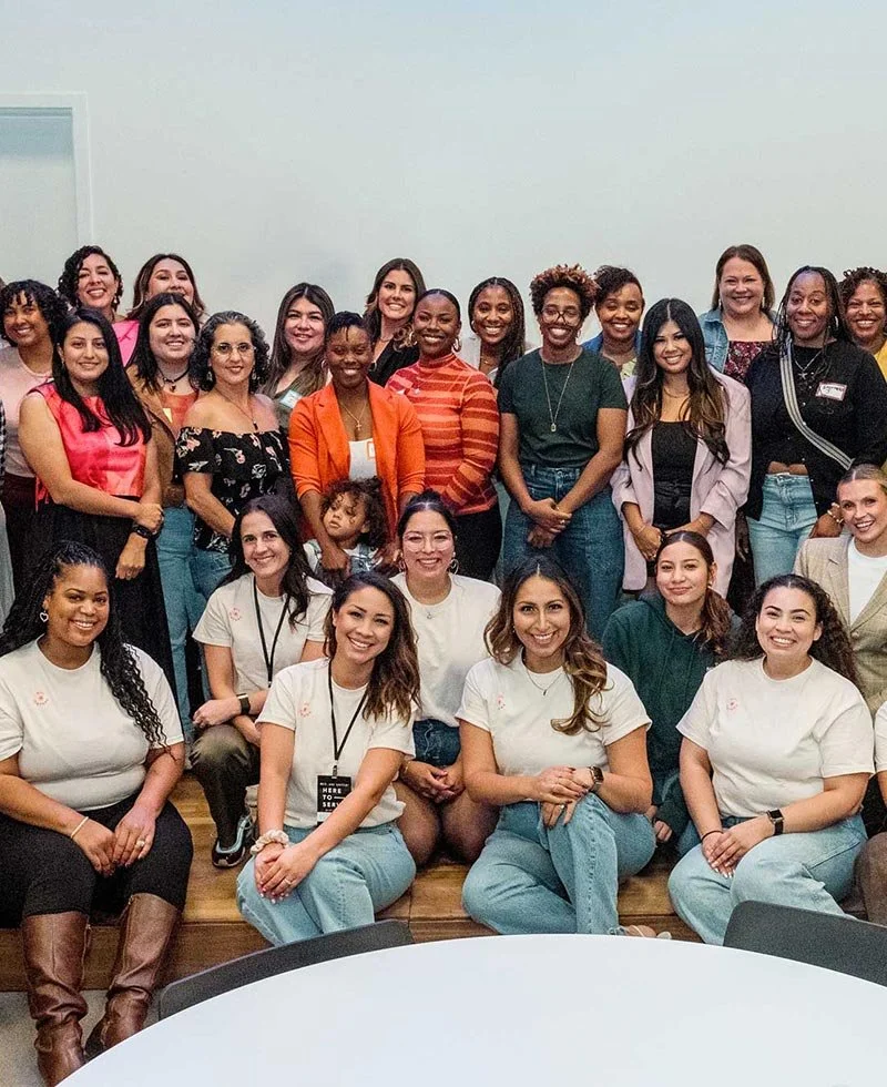 Group of women posing together in a room, some standing and some sitting in front, smiling at the camera.