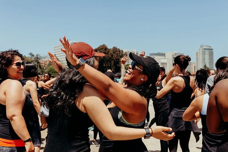 Two women smiling and embracing during an outdoor event with a crowd and city buildings in the background.