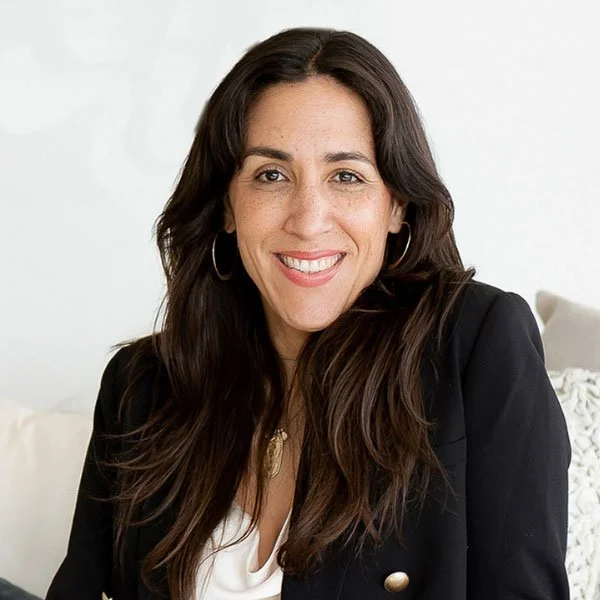 Woman with long brown hair smiling, wearing a black blazer and hoop earrings, sitting on a white couch in a bright room.