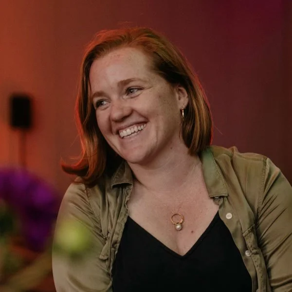 A woman with shoulder-length brown hair smiling, wearing a black top and an olive green blouse, in an indoor setting with a red background.