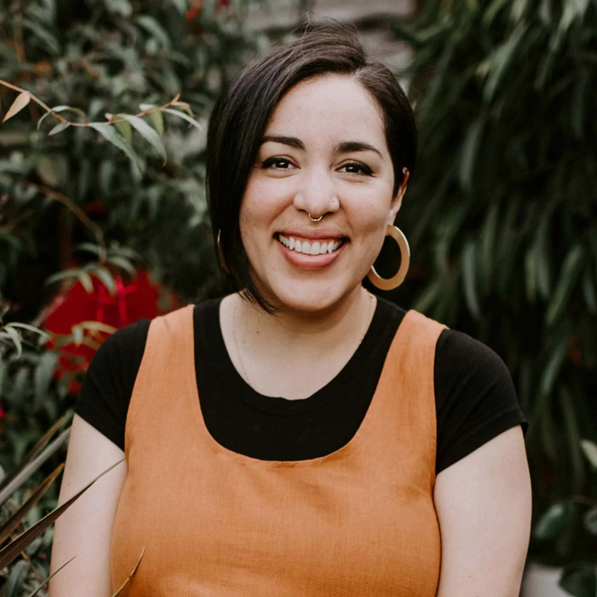 Portrait of a young woman with short dark hair and large hoop earrings, smiling in front of lush greenery.