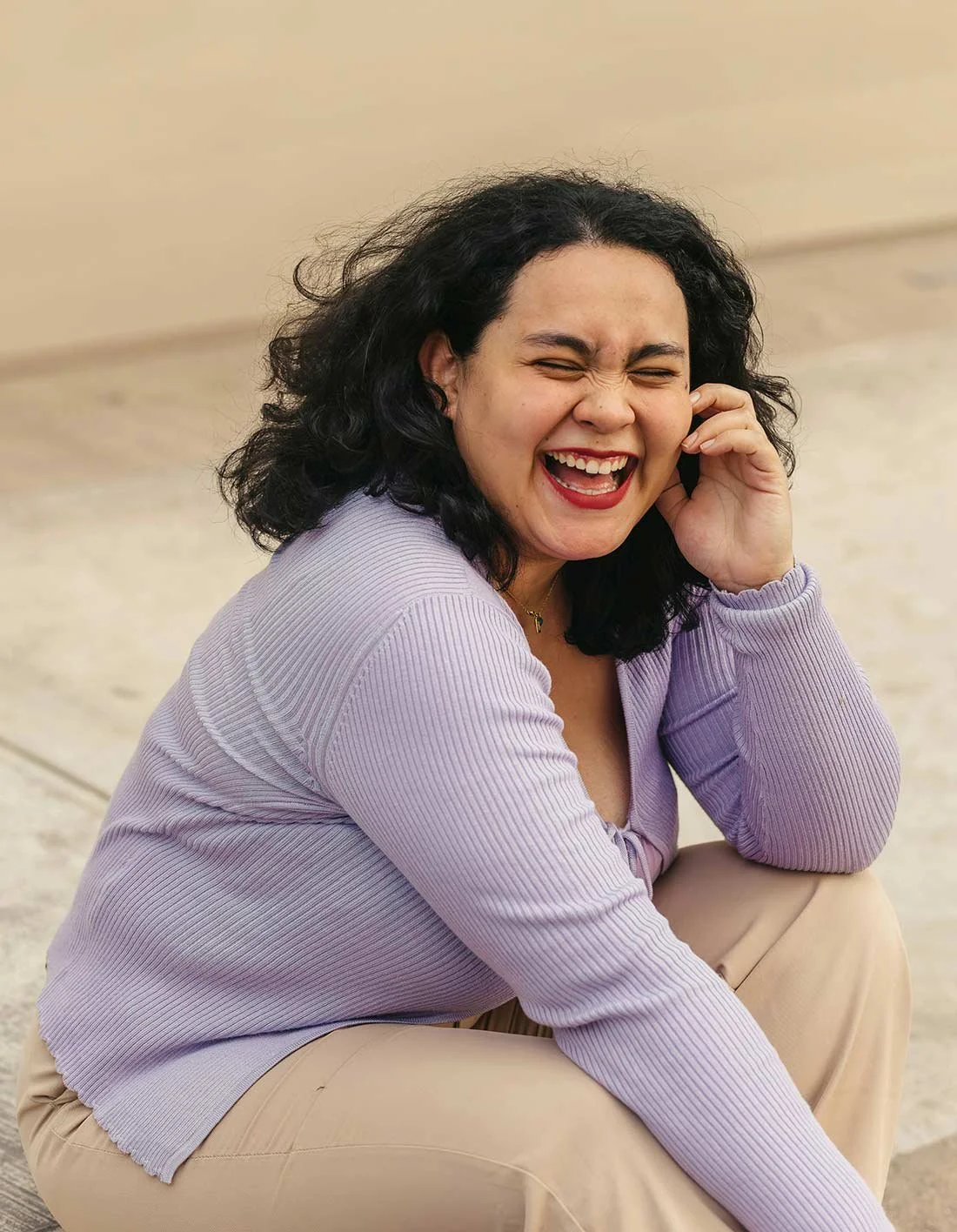 A woman with dark curly hair, wearing a lavender long sleeve shirt and beige pants, is sitting on the ground, laughing and talking on a phone.