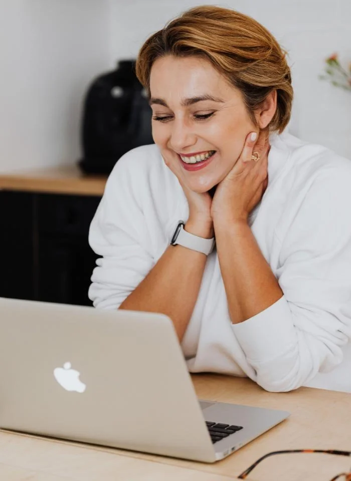 A woman in a white shirt smiling at her laptop, in a cheerful and relaxed moment.