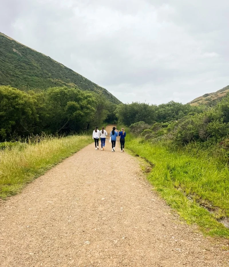 Four people walking together on a dirt trail in a green mountainous area with cloudy sky.