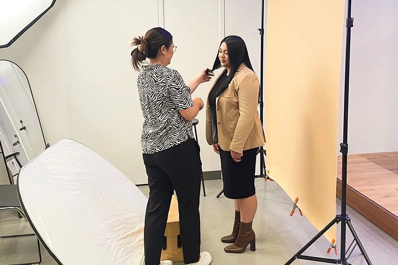 Two women in a photography studio, one adjusting the other's hair. The woman on the right is wearing a beige blazer and black skirt with brown boots, while the woman on the left is in a zebra print top and black pants. A beige backdrop and photograph
