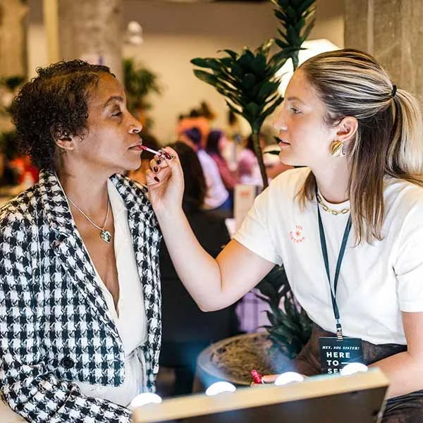 A makeup artist applies lipstick to a woman's lips at a makeup station in an indoor event space.