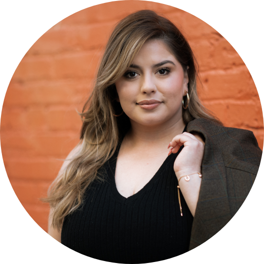 A young woman with wavy, shoulder-length hair standing in front of an orange brick wall, wearing a black V-neck top, hoop earrings, and a dark blazer draped over her shoulder.