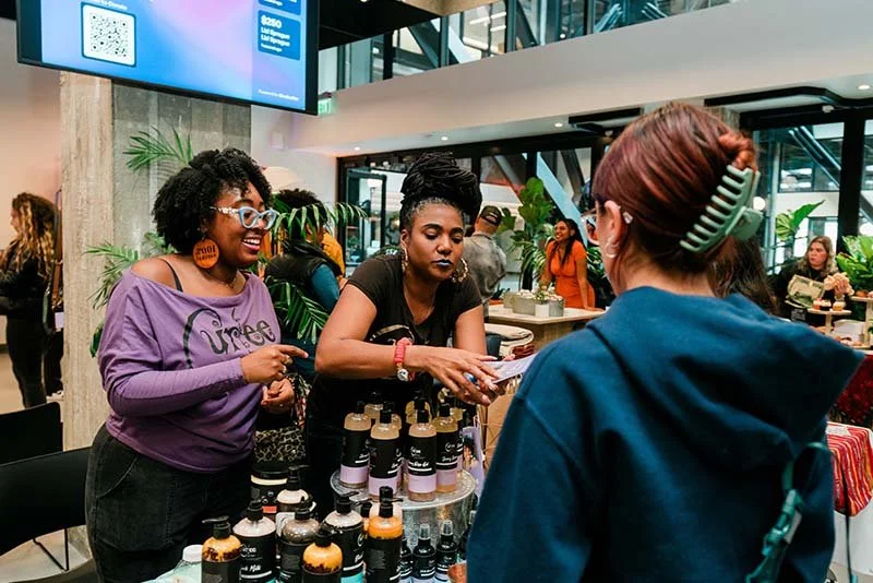 Two women at a booth with various skincare or cosmetic products, engaging with a customer in a busy indoor setting.