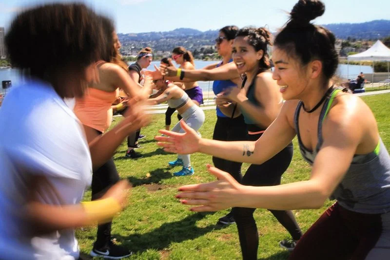 Group of women and girls participating in an outdoor team-building activity, running and passing hands on a grassy field with a view of distant hills and tents.