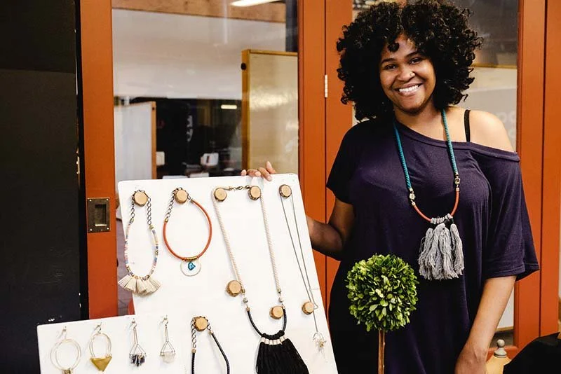 Woman standing next to jewelry display board with necklaces and earrings, smiling indoors.