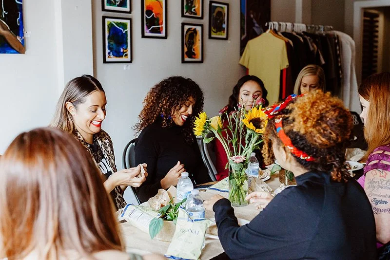 Group of women sitting around a table with sunflower centerpiece, engaged in conversation and laughter, in a room decorated with framed artwork and hanging clothes.