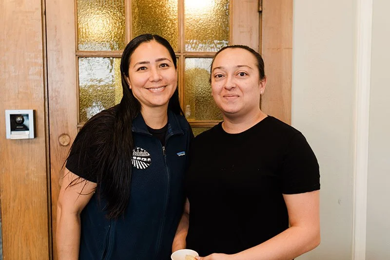 Two women standing indoors, smiling at the camera, with a wooden door and yellow glass window panes behind them.