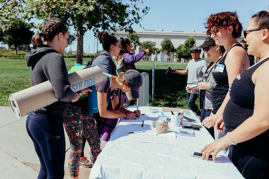 A group of women participating in an outdoor event, signing a document on a table, with others standing behind the table under a bright sunny day.
