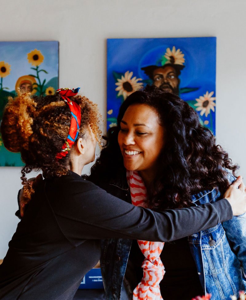 Two women are hugging and smiling at each other in an art gallery with colorful paintings of flowers and portraits in the background.
