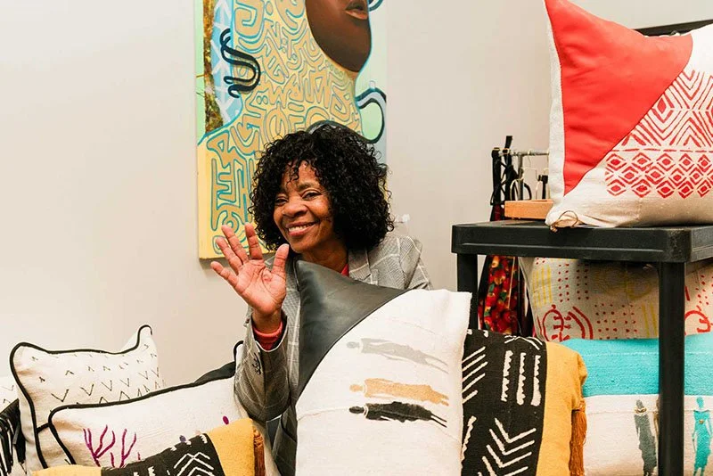 A woman with curly hair smiling and waving, surrounded by colorful pillows and artwork in a cozy indoor setting.