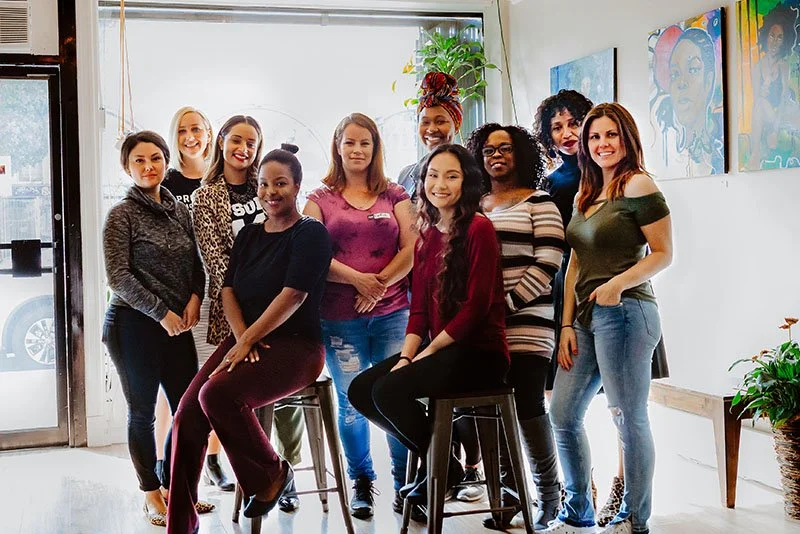 A diverse group of ten women posing together inside an art gallery or studio space, with artworks on the wall and plants in the background.