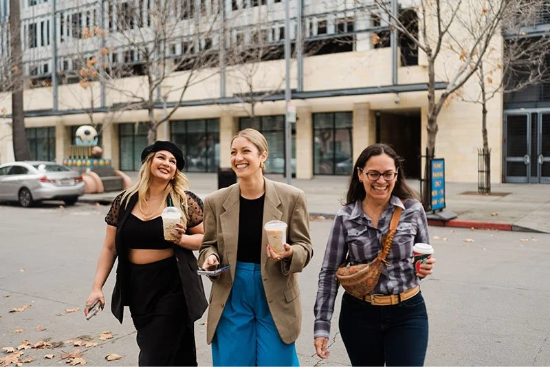 Three women walking on city street with trees and buildings in the background, smiling and holding coffee or drinks.