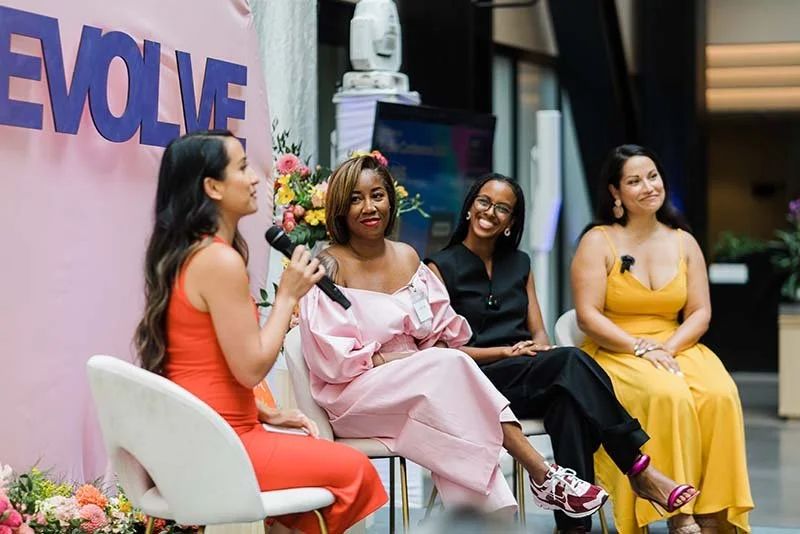 Four women sitting on stage in front of a pink backdrop with the word 'EVOLVE' on it, engaging in a panel discussion, with one holding a microphone and the others smiling.