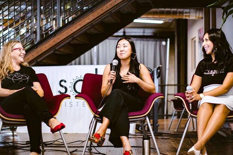 Three women sitting on stage in a panel discussion, with a staircase behind them. The woman in the middle is holding a microphone and gesturing with her hand, while the other two women are smiling.